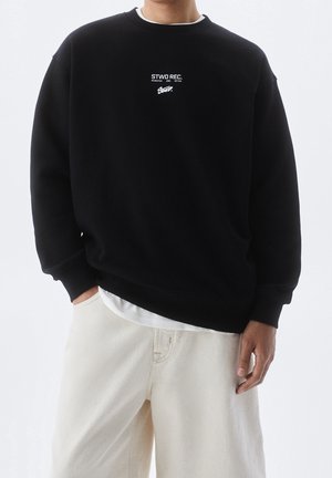 Sweatshirt noir avec col rond, arborant un logo en texte blanc sur la poitrine. Tissu doux avec des poignets côtelés, associé à un pantalon de couleur claire.