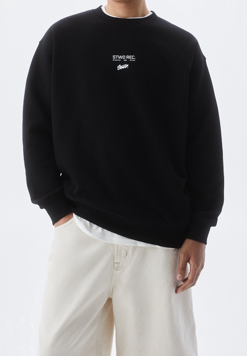 Černá mikina s kulatým výstřihem, se bílým textovým logem na hrudi. Měkká látka s pružnými manžetami, v kombinaci se světle barevnými kalhotami.