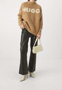 Sweater en tricot marron surdimensionné avec "HUGO" en blanc, pantalons en cuir noir évasés, sac à main crème et bottes à talons en imprimé serpent.