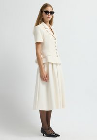 Femme portant un blazer crème à manches courtes avec des boutons et une jupe midi plissée, des collants noirs transparents, des escarpins T à talon noir et des lunettes de soleil en œil de chat.