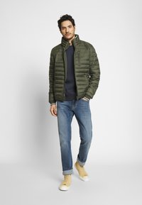 Veste rembourrée vert olive avec un col haut, fermeture éclair sur le devant et poches latérales, portée sur un pull bleu marine et associée à un jean bleu et des baskets jaunes.