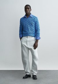 Homme debout, les mains dans les poches, portant un pull en maille bleu par-dessus une chemise rayée, un pantalon blanc et des chaussures noires sur un fond uni.