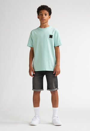 Lichtblauw katoenen T-shirt met een zwarte logopatch, gecombineerd met donker denim shorts en witte sneakers. De outfit heeft opgerolde zomen.