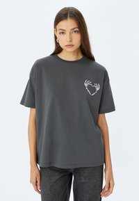 Camiseta de algodón gris de manga corta, con un diseño de corazón formado por una mano esquelética blanca en el lado izquierdo del pecho. Textura suave, corte relajado.