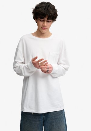 Ung mand med krøllet hår iført en hvid langærmet skjorte og blå jeans, mens han justerer ringe på fingrene, stående foran en ensfarvet baggrund.