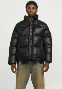Zwarte pufferjack met een hoge kraag, glanzende nylon stof, gestikte delen, twee voorzakken en een relaxed fit, gedragen over een geruite overhemd.