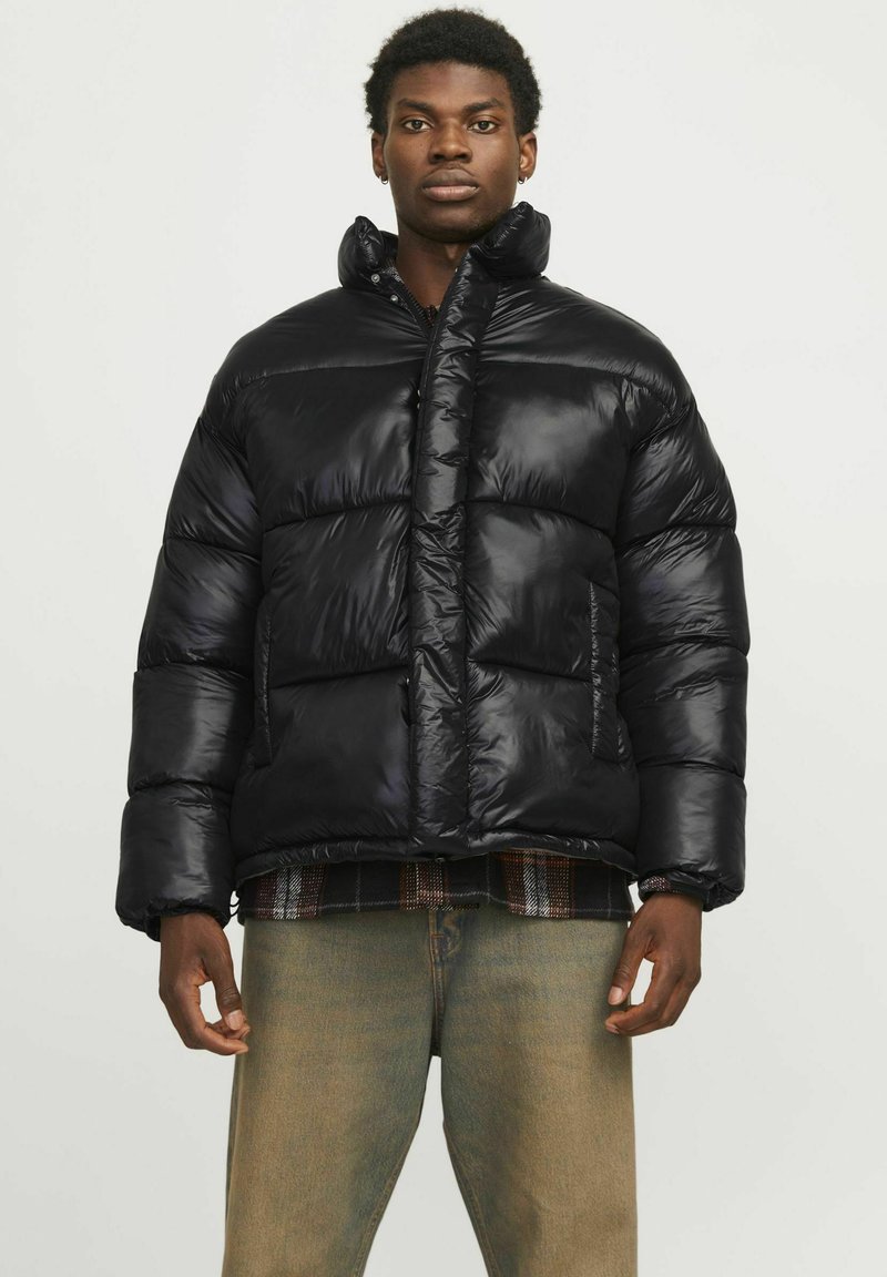 Zwarte pufferjack met een hoge kraag, glanzende nylon stof, gestikte delen, twee voorzakken en een relaxed fit, gedragen over een geruite overhemd.