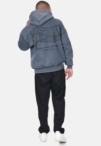 Übergroßer grauer Kapuzenpullover mit einem großen schwarzen Logo auf der Rückseite, gerippten Bündchen und einer Kordelzugkapuze. Kombiniert mit schwarzen Hosen und weißen Sneakern.