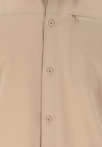 Chemise beige à boutons avec une petite poche zippée sur le côté gauche de la poitrine et quatre boutons visibles alignés verticalement.