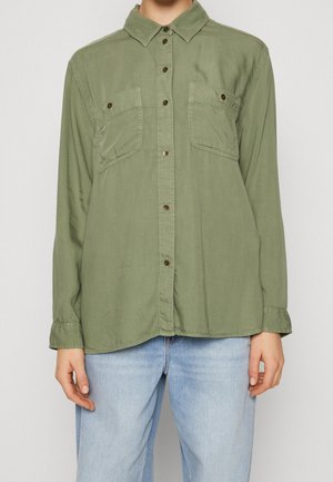 Personne portant une chemise boutonnée vert olive avec deux poches poitrine boutonnées et un jean bleu clair sur un fond uni.