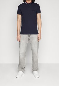 Man som bär en marinblå kortärmad pikétröja, ljusgrå jeans och vita sneakers, stående mot en enkel ljus bakgrund.