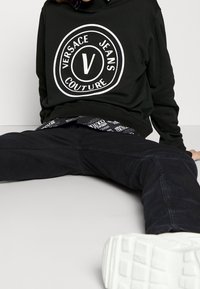 Svart sweatshirt med stor vit cirkulär logotyp med texten "Versace Jeans Couture". Kombinerad med svarta jeans och vita sneakers.