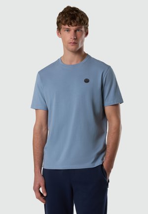 Uomo con capelli ricci che indossa una T-shirt a maniche corte azzurro chiaro con un piccolo logo rotondo nero sul petto e pantaloni scuri, in piedi con una mano in tasca.