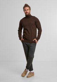 Brauner Rollkragenpullover aus weichem Material, mit langen Ärmeln und einer taillierten Form, kombiniert mit grauen Hosen und hellbraunen Schuhen.