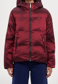 Veste rouge matelassée à finition brillante, dotée d'une capuche, d'une fermeture éclair avant et de deux poches latérales zippées. Structurée avec des coutures horizontales.