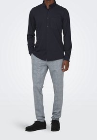 Granatowa koszula zapinana na guziki z kieszenią na piersi, zestawiona z jasnoszarymi, teksturowanymi spodniami i czarnymi butami, w pozycji stojącej na neutralnym tle.