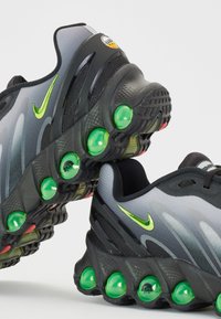 Czarne i szare buty sportowe z zielonymi słuchawkami AirPods, charakteryzujące się smukłym designem, teksturowaną cholewką oraz kontrastującymi żółtymi akcentami.