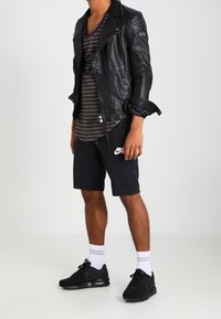 Personne portant une veste en cuir noir, une chemise rayée, un short Nike noir, des chaussettes blanches et des baskets noires, se tenant devant un fond uni.