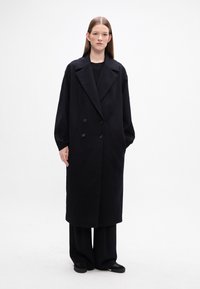 Abrigo negro de doble botonadura con solapa ancha y mangas largas, hecho de una tela suave con una textura lisa. Se lleva sobre pantalones holgados negros.
