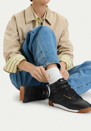 Person sitzt mit einem angewinkelten Knie und richtet weiße Socken, trägt schwarze Reebok-Sneakers, blaue Jeans, eine beige Jacke und ein gelb gestreiftes Hemd.