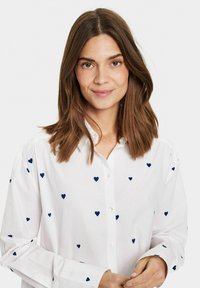 White button-up shirt featuring small navy heart patterns, long sleeves, and a classic collar design. Smooth texture and tailored fit.