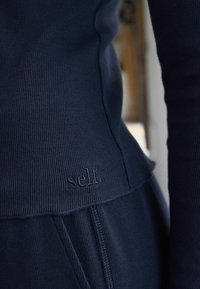 Tengerészkék, bordázott, hosszú ujjas felső, finoman dombornyomott "self." logóval a szegélyén, fitted szabással és puha textúrával.