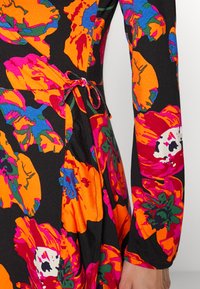 Vestido de manga longa com padrões florais bold e multicoloridos sobre um fundo preto, com detalhe de cintura atada e tecido macio e texturizado.
