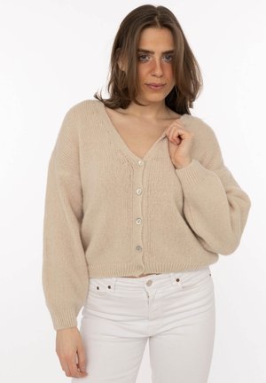Frau trägt eine beige, geknöpfte Strickjacke mit langen Ärmeln und weiße Hose, hält den Kragen der Strickjacke mit einer Hand.