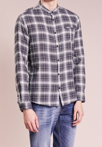 Szara i biała koszula w kratkę z lekkiego materiału, z kołnierzykiem mandaryńskim, przednią kieszenią i czarnymi guzikami. Noszona z niebieskimi dżinsami.