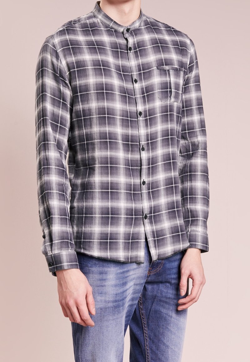 Szara i biała koszula w kratkę z lekkiego materiału, z kołnierzykiem mandaryńskim, przednią kieszenią i czarnymi guzikami. Noszona z niebieskimi dżinsami.