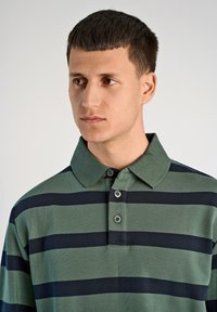 Polo w zielono-granatowe paski z kołnierzykiem, trzema guzikami oraz prążkowaną fakturą, prezentowany na modelu z krótkimi włosami.