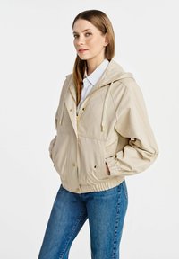 Beżowa kurtka z kapturem wykonana z gładkiego materiału, z pikowanym wzorem, złotym wykończeniem, elastycznymi mankietami i ściągaczem w pasie.