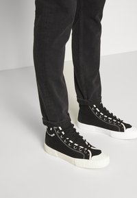 Zapatillas deportivas altas negras con suela de goma blanca, ojales acentuados y cordones blancos en contraste sobre una parte superior de lona. Jeans negros con el dobladillo vuelto.