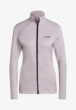 Kobieta ubrana w jasnoróżową kurtkę zapinaną na zamek błyskawiczny, ciemne legginsy i czarne sportowe buty Adidas, stojąca z jedną ręką w kieszeni.
