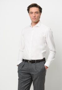 Camicia bianca a maniche lunghe con colletto classico, abbinata a pantaloni grigi con texture e una cintura in pelle marrone con fibbia argentata.