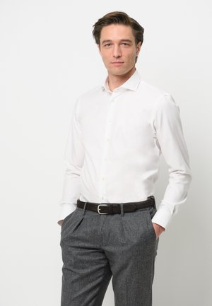 Chemise blanche à manches longues avec un col classique, associée à un pantalon gris texturé et une ceinture en cuir marron avec une boucle en argent.