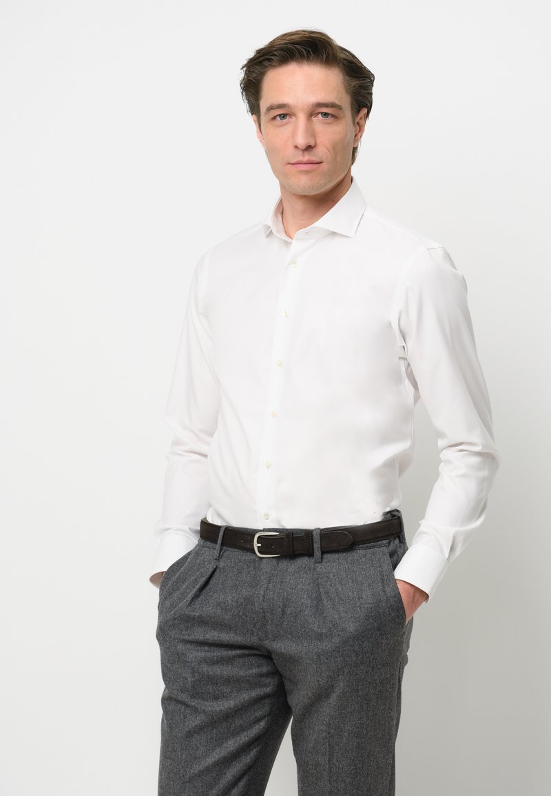 Camicia bianca a maniche lunghe con colletto classico, abbinata a pantaloni grigi con texture e una cintura in pelle marrone con fibbia argentata.