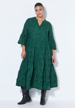 Frau mit kurzen Haaren, die ein dunkelgrünes, langärmeliges, besticktes Kleid und schwarze Stiefel bis zur Mitte der Wade trägt, steht vor einem weißen Hintergrund.