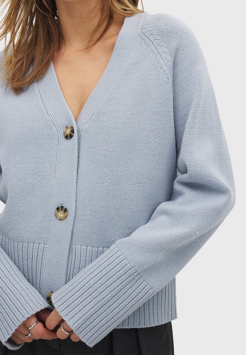 Femme portant un cardigan en maille bleu clair avec trois grands boutons et des poignets côtelés, montrée de la poitrine à la taille.