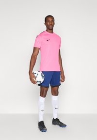 Ποδοσφαιρική φανέλα Nike ροζ, ναυτικές σορτς, λευκές κάλτσες, μαύρα ποδοσφαιρικά παπούτσια; αθλητής κρατάει μια σχεδιασμένη μπάλα ποδοσφαίρου, στέκεται σε ουδέτερο φόντο.