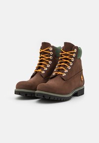 Bruine leren wandelschoenen met oranje veters, groene kraag en een robuuste zwarte zool met een onderscheidend logo aan de zijkant.