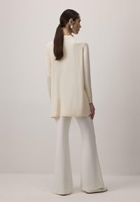 Krém színű, hosszú ujjú blúz, laza fazonnal, krém színű, testhez simuló, harangszárú nadrággal párosítva, amely sima, puha textúrával rendelkezik.
