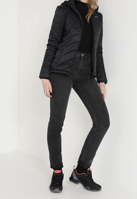 Veste matelassée noire avec capuche, chemise noire à manches longues, jean skinny foncé et chaussures de randonnée noires avec des accents roses.