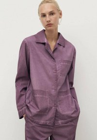 Purple button-up shirt with a collar, featuring two front pockets and a relaxed fit. Smooth texture with a slightly crinkled appearance.