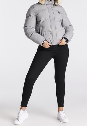 Persona con chaqueta acolchada gris claro con capucha de pelo, pantalones pitillo negros y zapatillas blancas, de pie con las piernas separadas y las manos cerca de la cintura.