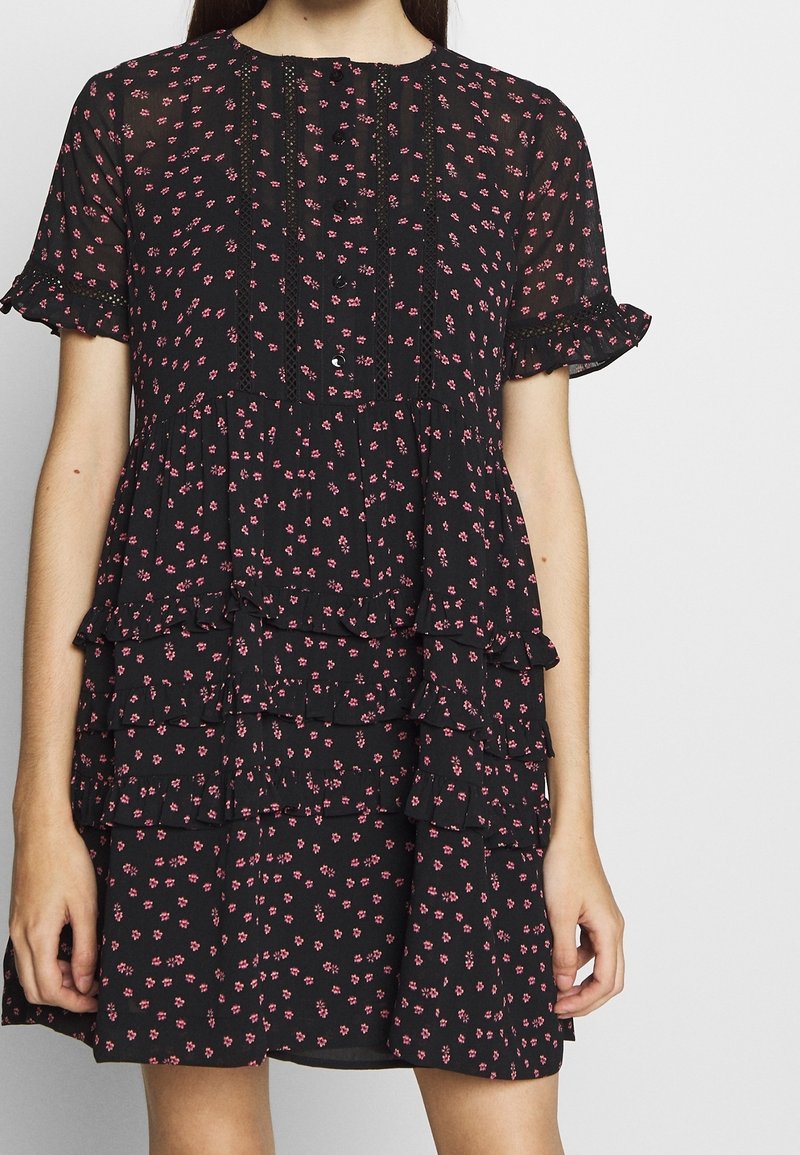 Femme portant une robe noire à petits motifs floraux roses, manches courtes à volants, boutons sur le devant et jupe à volants superposés.
