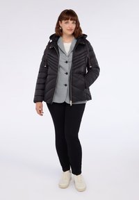 Kvinna klädd i en svart hooded puffer-jacka över en rutmönstrad kavaj, svarta byxor och vita sneakers, stående framför en vit bakgrund.