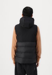 Svart polstret vest med høy krage og hette, med en matt overflate og horisontalt kviltet design, brukt over en beige langermet skjorte.