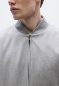 Szürke bomber dzseki cipzáros záródással, kontrasztos szürke kötött gallérral és sima, texturált anyaggal. Tiszta, modern design.