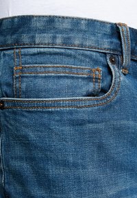 Primer plano de un bolsillo de jeans de mezclilla azul con costuras naranjas y remaches negros en la cintura y el costado.